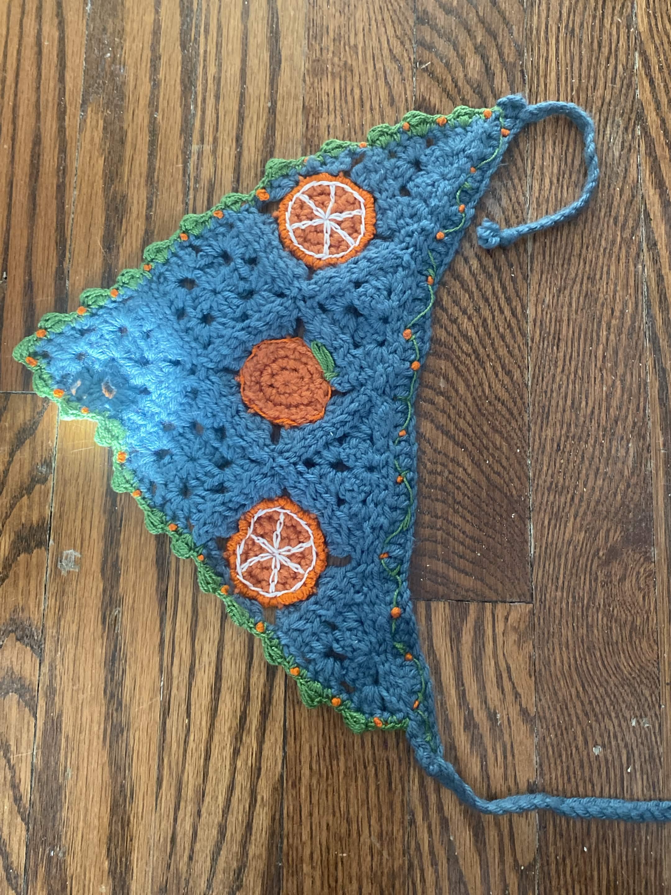 dusty blue crochet triangle headscarf. two tangering slices and one tangerine are aligned on the wide end. vine with orange spheres boarders the opposing sides.